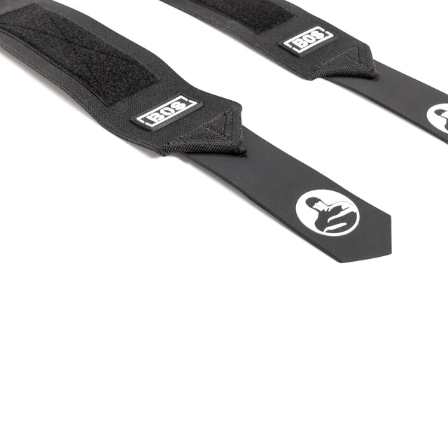 Two black Mighty Wrist Wraps by Bells of Steel, featuring Velcro fastenings and a white stylized helmet logo near the pointed end, are laid out diagonally on a white surface in competition style.