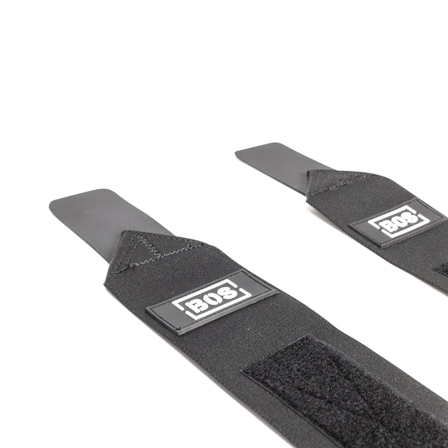 Two black Mighty Wrist Wraps by Bells of Steel, each with hook-and-loop fasteners and rectangular BOS labels, featuring flat stiff ends for wrist support—ideal for powerlifting—laid out on a white background.
