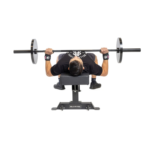 A person lies on a bench, gripping a barbell and wearing Bells of Steel Mighty Wrist Wraps—an essential accessory for powerlifters—while getting ready to perform a bench press. The top-down view shows the barbell and bench clearly.