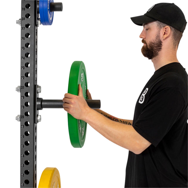 A man in a black t-shirt and cap places a green weight plate onto the Bells of Steel Bolt-On Plate Peg Sets, which also hold blue and yellow plates for organized storage on the power rack.