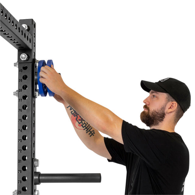 A man in a black cap and t-shirt loads blue Olympic plates onto Bells of Steel Bolt-On Plate Pegs, which are mounted on a metal power rack against a white background.