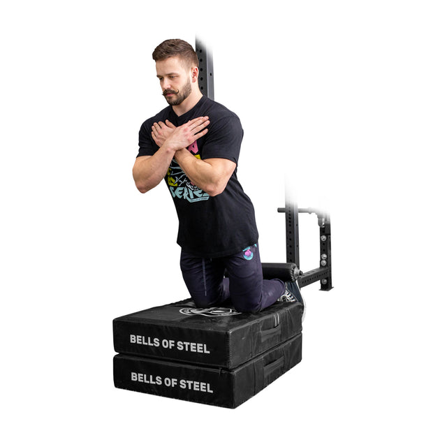 A man kneels on two stacked Bells of Steel Hydra Nordic Curl Packages with arms crossed, demonstrating hamstring curls against a white background.
