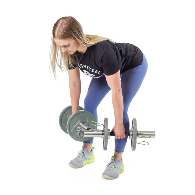 A woman in a black t-shirt and blue leggings performs a bent-over row with Bells of Steel Loadable Dumbbell Handles, gray sneakers on her feet. She looks down, keeps her back straight, and the white background highlights home gym equipment.