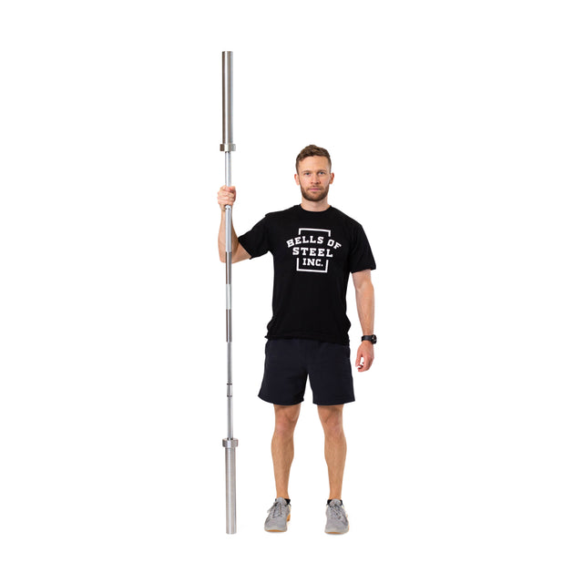 A man in a black Bells of Steel t-shirt and shorts stands on a white background, holding the Bells of Steel Olympic Weightlifting Barbell with needle bearings vertically in one hand.