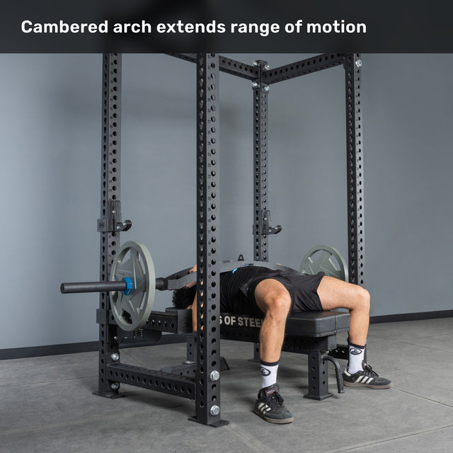 Lying on a bench with an arched back, a person grips the Bells of Steel Arch Nemesis Swiss Bar in a power rack. Text above reads: "Cambered arch extends range of motion." This specialty bar boosts pressing performance.