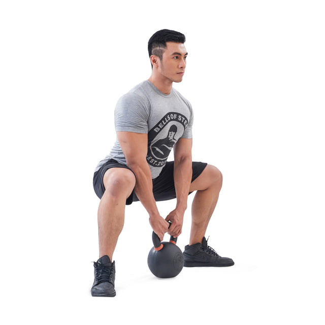 A man in a gray t-shirt and black shorts demonstrates strength training with a Bells of Steel Powder Coated Kettlebell, performing a squat while holding it with both hands against a white background.