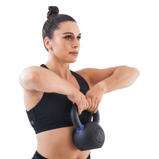 A woman in athletic wear showcases strength training, performing an upright row to chest level with both hands using a Bells of Steel Powder Coated Kettlebell, set against a white background.