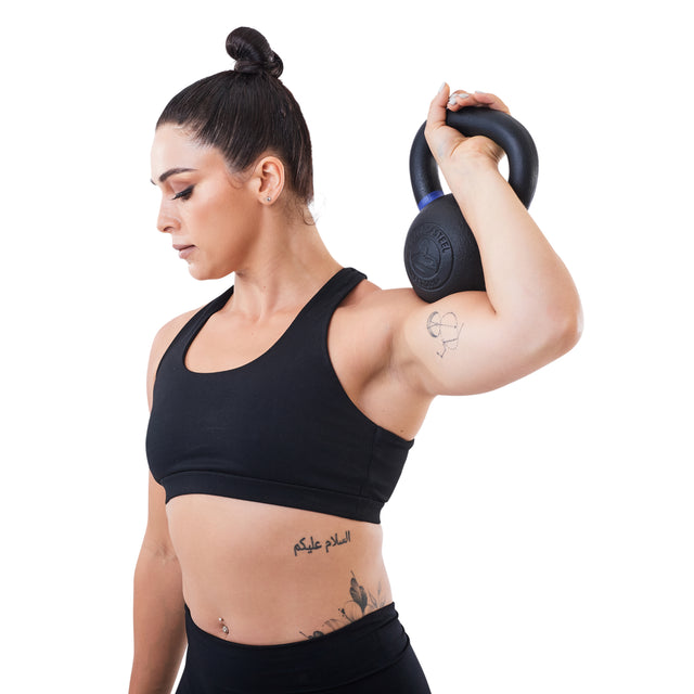 A tattooed woman with her hair in a bun holds a Bells of Steel Powder Coated Kettlebell on her shoulder, wearing a black sports bra and leggings, ready for functional fitness against a white background.