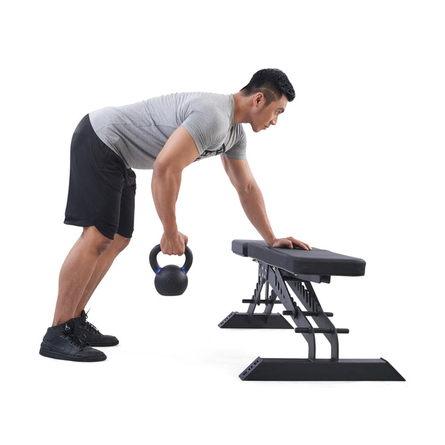 A man in a gray t-shirt and black shorts demonstrates strength training by performing a bent-over row with a Bells of Steel Powder Coated Kettlebell, using a workout bench for support against a white background.