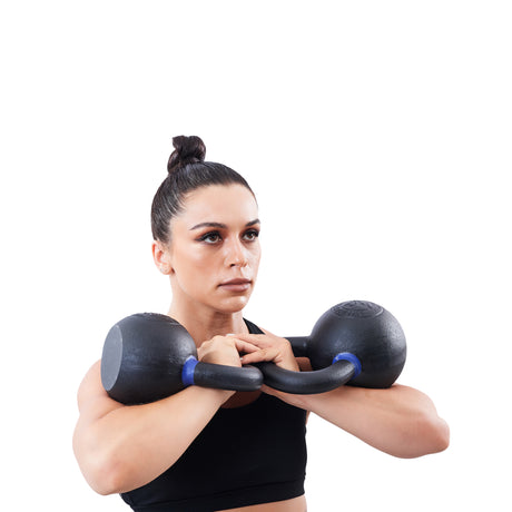 A woman with dark hair in a bun holds a Bells of Steel Powder Coated Kettlebell at chest level. Dressed in a black sports bra, she looks forward with focus against a white background.
