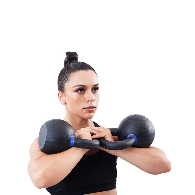 A woman with dark hair in a bun holds a Bells of Steel Powder Coated Kettlebell at chest level. Dressed in a black sports bra, she looks forward with focus against a white background.