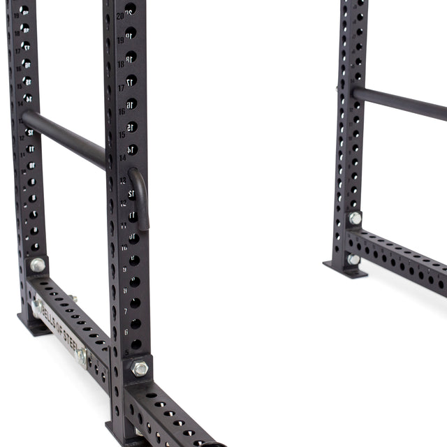 Close-up of a Bells of Steel black metal power rack featuring numbered holes, two vertical posts, and horizontal support bars. Included are Pin Pipe Safeties for added protection during squats and bench presses.