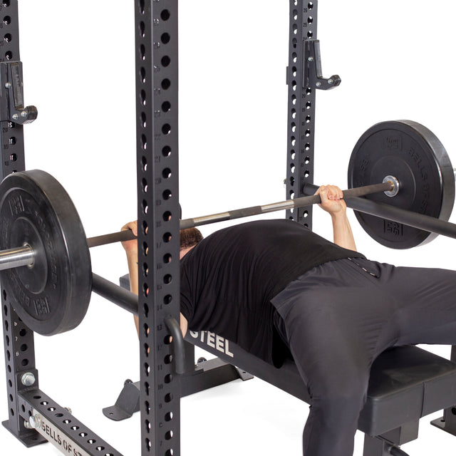 A person in black workout clothes lies on a bench, gripping a barbell and preparing to bench press inside a power rack equipped with Bells of Steel Pin Pipe Safeties for added protection.