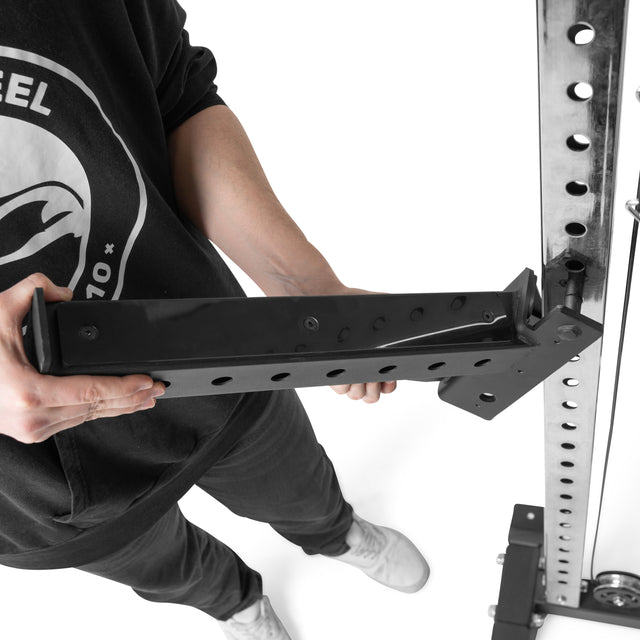 Wearing a black hoodie, a person is precisely installing a metal bracket on the Bells of Steel Functional Trainer Cable Tower. They hold the bracket against the tower rack with both hands. The white background emphasizes their focused effort.