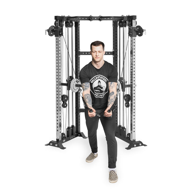 A tattooed individual in a black t-shirt with a white logo, black pants, and checkered shoes uses the Bells of Steel Functional Trainer Cable Tower for a cable crossover workout, standing out against the plain white background.