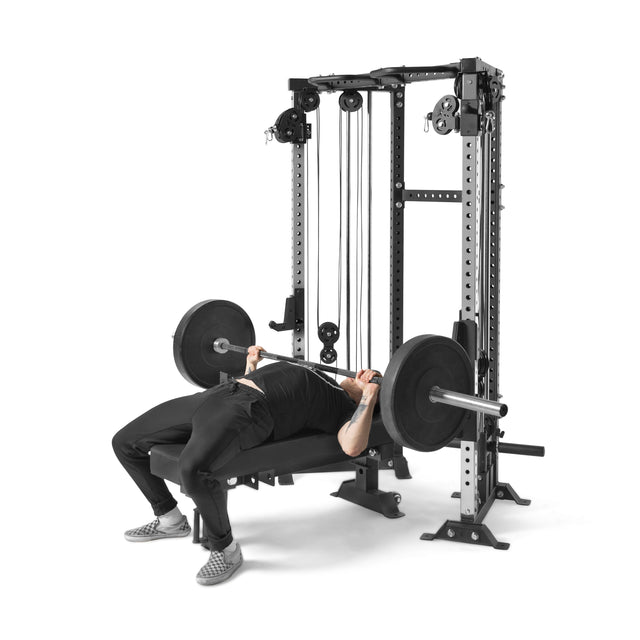 Wearing black athletic clothes and checkered shoes, a person bench presses with a barbell on the Smith machine of the Bells of Steel Functional Trainer Cable Tower, effortlessly using its adjustable cables and weight stacks.