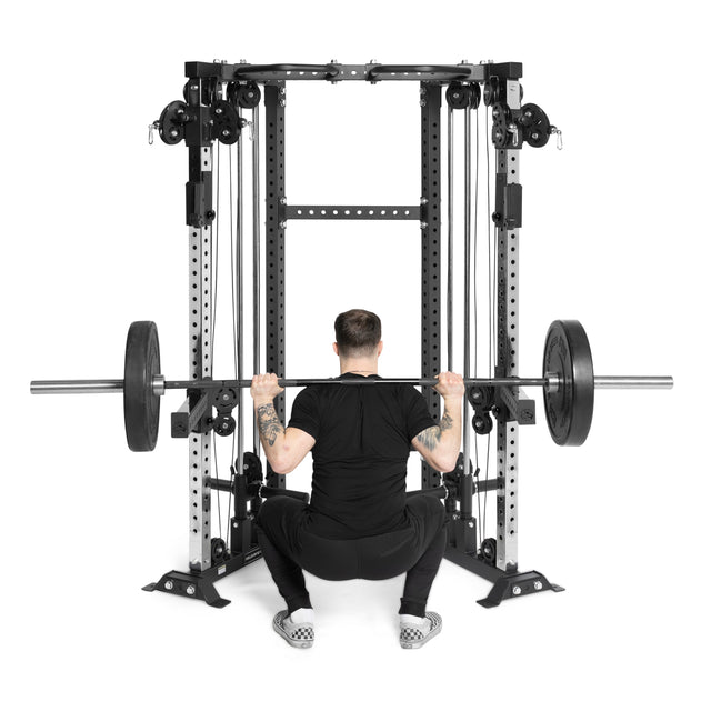 A person in black workout clothes and checkered shoes is squatting with a barbell in a gym equipped with Bells of Steel Functional Trainer Cable Towers and weight stacks, perfect for serious lifts and versatile workouts.