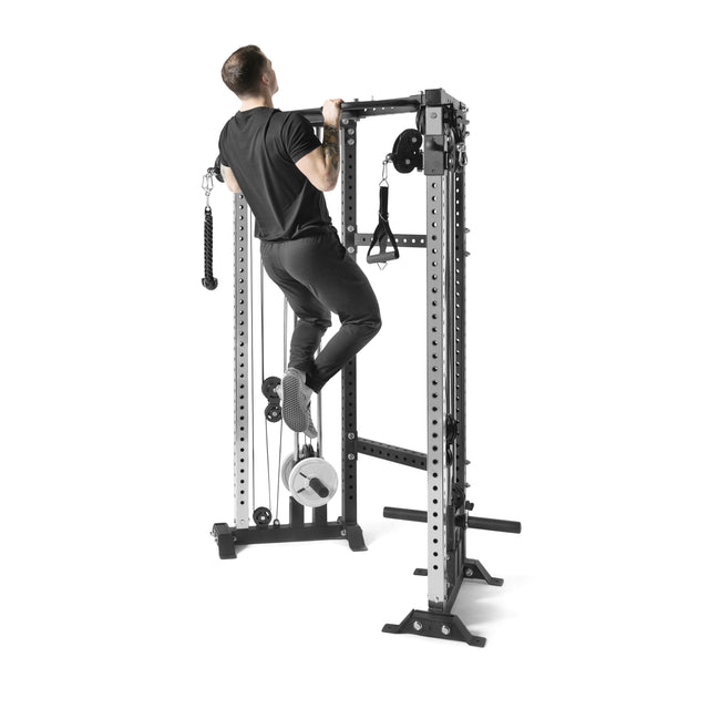 A person uses the Bells of Steel Functional Trainer Cable Tower for pull-ups on a gym power rack, with various attachments and weights, set against a plain white background.
