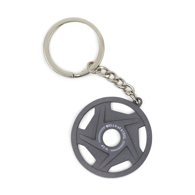 The Plate Keychain by Bells of Steel features a silver ring and a round, dark gray metal charm resembling a 0.5 lb weight plate, engraved with "BELLS OF STEEL" and "0.5 LB" in white.