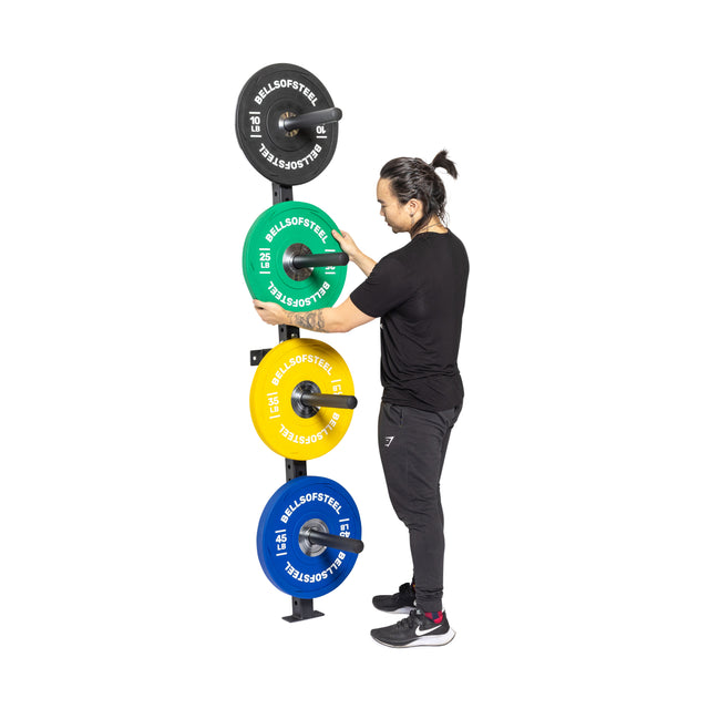 A person in black workout gear places a green weight plate on the Bells of Steel Wall-Mounted Plate Storage Rack, neatly organizing colorful plates and demonstrating efficient storage against a white backdrop.
