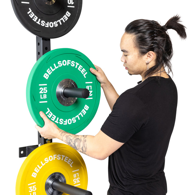 A person with a black shirt and ponytail places a green 25 lb Bells of Steel plate onto a Wall-Mounted Plate Storage Rack, which also holds yellow and black plates for efficient home gym storage.