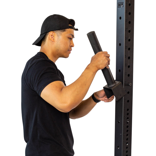 A person in a black t-shirt and backward cap uses a black Pin Plate Peg from Bells of Steel to install rack-mountable plate pegs on a metal fitness rack with numbered holes—ideal for Olympic weight plate storage.