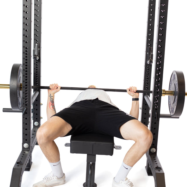 A person bench presses on a gym bench with a barbell, while Bells of Steel Pin Pipe Safeties on the power rack provide extra protection during the lift.