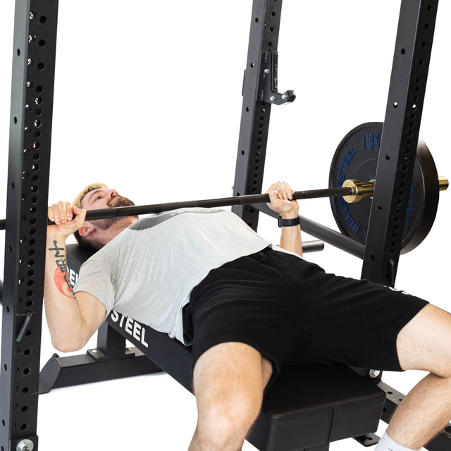 A man in a grey t-shirt and black shorts bench presses a barbell inside a power rack equipped with Bells of Steel Pin Pipe Safeties for protection, gripping the bar with both hands against a white background.