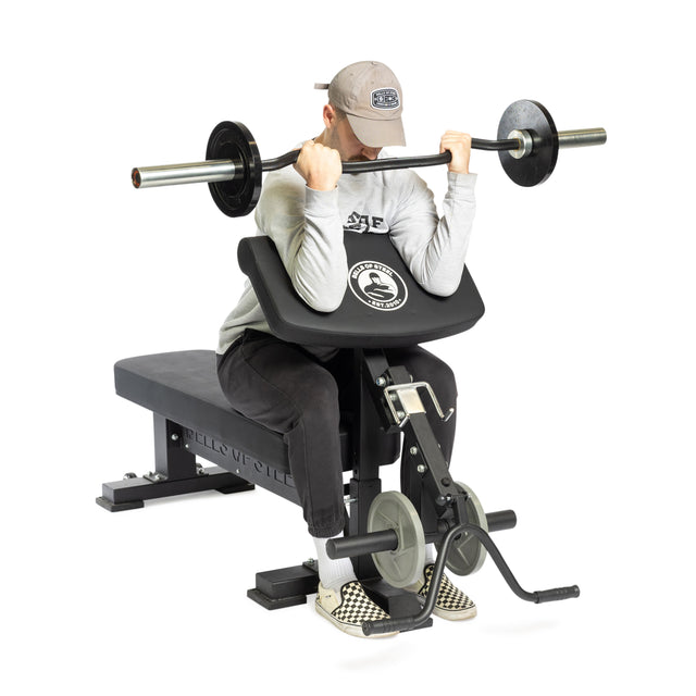 A person in a cap and casual clothes is seated, performing a barbell preacher curl using Bells of Steel Bench Attachments on a bench, isolated on a white background.