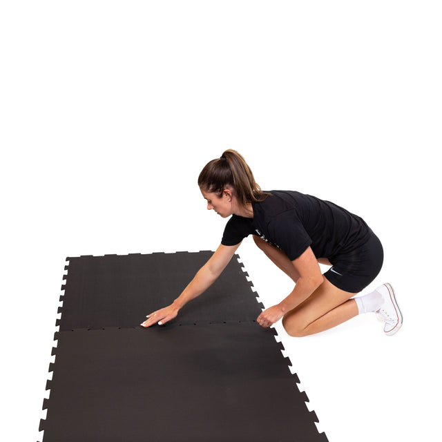 Wearing athletic gear, a person kneels on the floor connecting Bells of Steel Black Puzzle Mats against a white background.