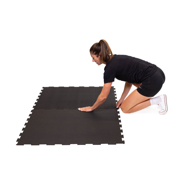 A woman in athletic wear kneels on the floor, connecting two Bells of Steel Black Puzzle Mats together against a white background.