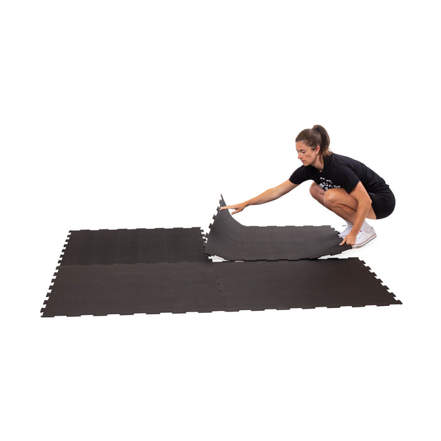 A person in athletic wear kneels assembling Bells of Steel Black Puzzle Mats—interlocking pieces ideal for gym flooring—on a white background.