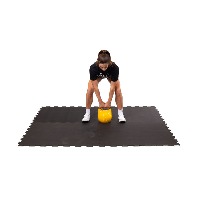 A woman in a black shirt, shorts, and white shoes stands on Bells of Steel Black Puzzle Mat, holding a yellow kettlebell with both hands as she prepares to lift it.