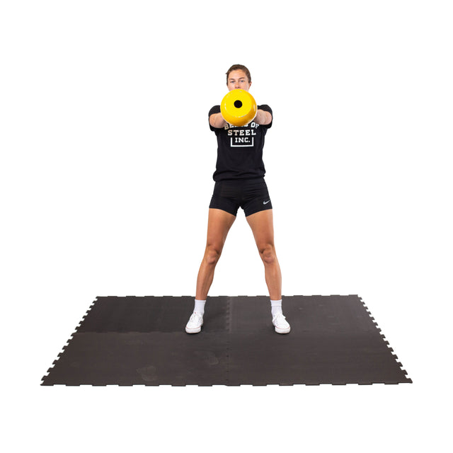 A woman stands on a Bells of Steel Black Puzzle Mat atop gym flooring, holding a yellow cylindrical weight at chest height with arms extended. She wears a black T-shirt, black shorts, and white sneakers.