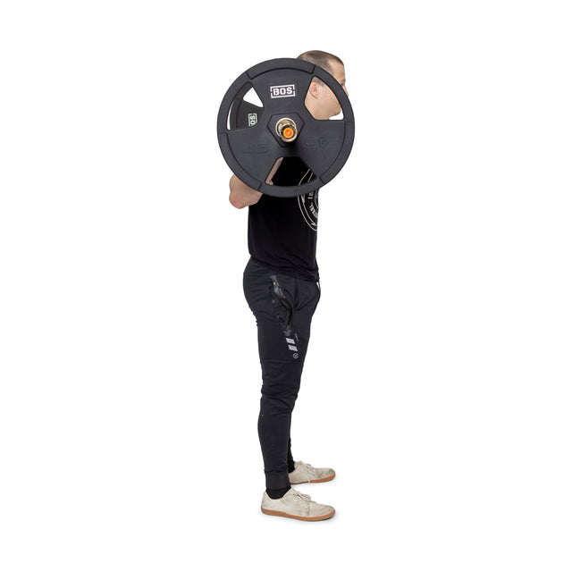 A person stands sideways holding Bells of Steel's Rubber Coated Iron Weight Plate Sets on their back. They're wearing a black t-shirt, dark joggers, and white sneakers, with the image focusing on their balanced weightlifting posture.