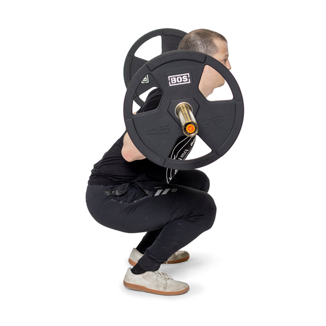 In black athletic wear, a person squats with Bells of Steel's Rubber Coated Iron Weight Plate Sets on their shoulders. The barbell, with rubber-coated plates and stainless steel hubs, looks sleek. They maintain a straight back and look forward as they squat low.