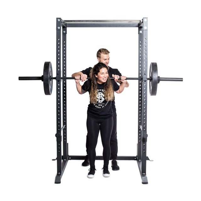 A woman prepares to squat with a barbell in the Bells of Steel Residential Power Rack (2.3" x 2.3", ⅝" HOLES) as a man spots her. Both, dressed in black athletic wear and smiling, stand against a white background—ideal for any home gym.