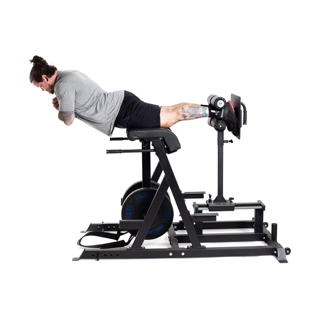 A tattooed person with a man bun uses the Reverse Hammer 2 In 1 GHD and Reverse Back Extension by Bells of Steel for hyperextension exercises. Wearing a gray shirt, black shorts, and colorful socks, they lean forward with arms crossed to target their posterior chain.