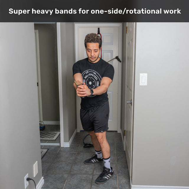 Wearing gym clothes, a man stands in a hallway performing an arm rotation exercise with Ripcords Premium Resistance Bands by Bells of Steel, using the super heavy band equipped with sturdy rubber handles for effective one-sided and rotational training, as shown in the text above.