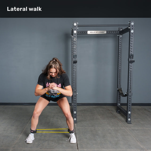 A woman performs a lateral walk exercise at the gym, squatting slightly and stepping sideways with Bells of Steel Human Trainer - Leg Cords around her legs for added stability. “Lateral walk” is written at the top left.