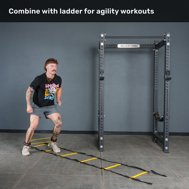 A person performs agility drills on a yellow ladder laid out on the gym floor, enhancing the workout with Bells of Steel Human Trainer - Leg Cords for increased resistance. A squat rack is visible nearby. Text reads, “Combine with ladder for agility workouts.”.