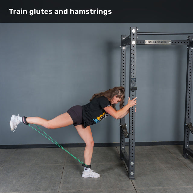A person performs a resistance band kickback, attaching Bells of Steel Human Trainer - Leg Cords to their ankle and a squat rack to target glutes and hamstrings. Leaning forward and holding the rack for support, they extend their leg back. Text reads, “Train glutes and hamstrings.”.