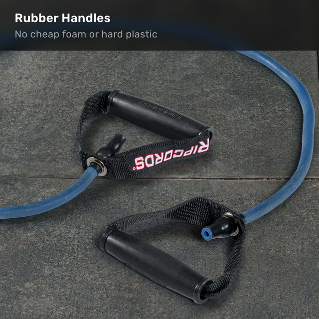 A blue Human Trainer - Ripcords resistance band by Bells of Steel, featuring black rubber grip handles and “RIPCORDS” in red on the strap, lies on a dark stone floor. Text above states: “Rubber Handles. No cheap foam or hard plastic.”.