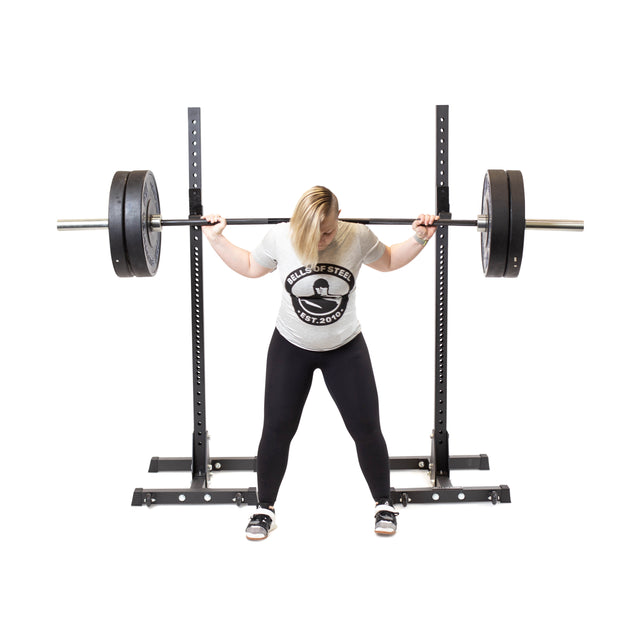 In their home gym, a person grips a barbell on Bells of Steel Squat Stands (2.3" x 2.3", ⅝" holes), wearing a light shirt, black leggings, and sneakers, head down in preparation to lift.