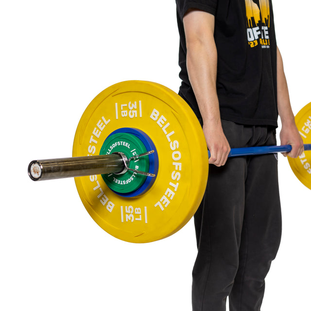 In black pants and shirt, a person holds a polished chrome barbell with large yellow "BELLS OF STEEL" 35 lb plates secured by Olympic barbell sleeves and grips the product from "Bells of Steel.