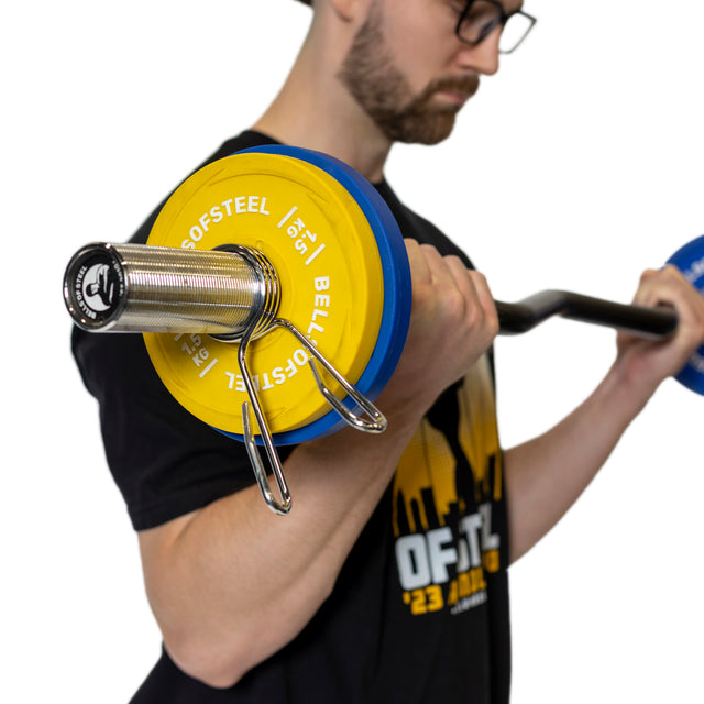 A man in glasses and a black t-shirt performs a bicep curl with a barbell featuring yellow and blue weight plates, secured by Bells of Steel Spring Collars, against a white background.