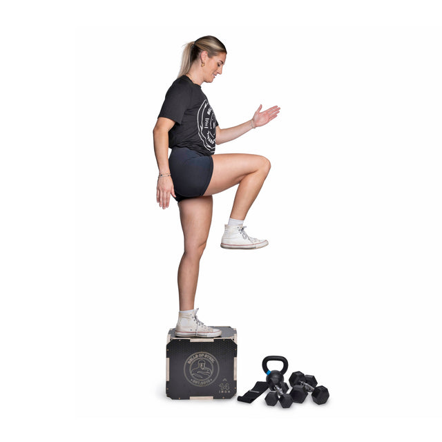 A person in athletic wear steps onto a workout box, lifting one knee while balancing. Bells of Steel Strength Circuit Starter Kits are on the floor beside the box. The plain white background sets the scene for a focused home workout.
