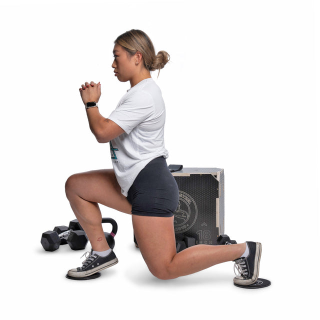 A woman in athletic wear lunges with clasped hands. Bells of Steel Strength Circuit Starter Kits, including dumbbells and a plyo box—perfect for home workouts—are visible on a white surface in the background.