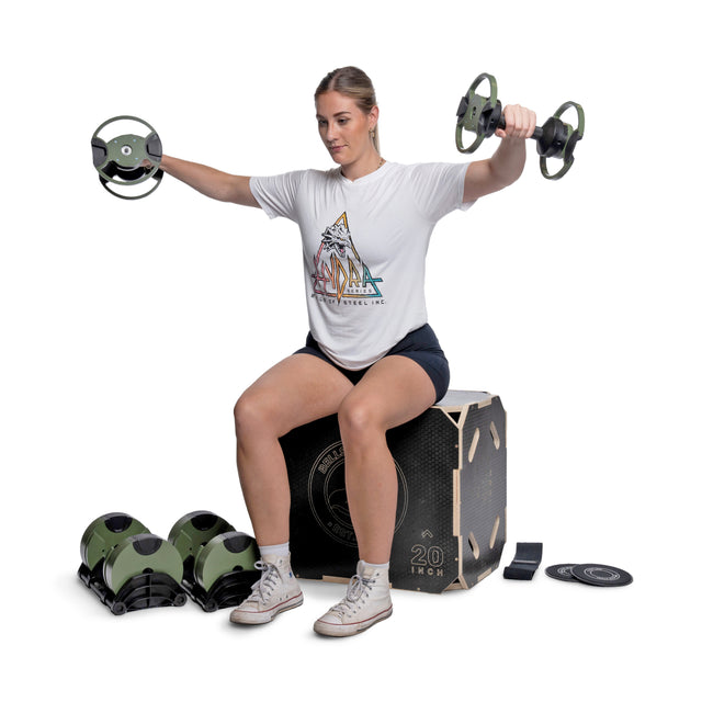 A woman in workout clothes sits on a plyometric box, lifting dumbbells from the Bells of Steel Strength Circuit Starter Kits. Balance trainers and resistance bands are nearby, creating an ideal home workout setup against a white background.