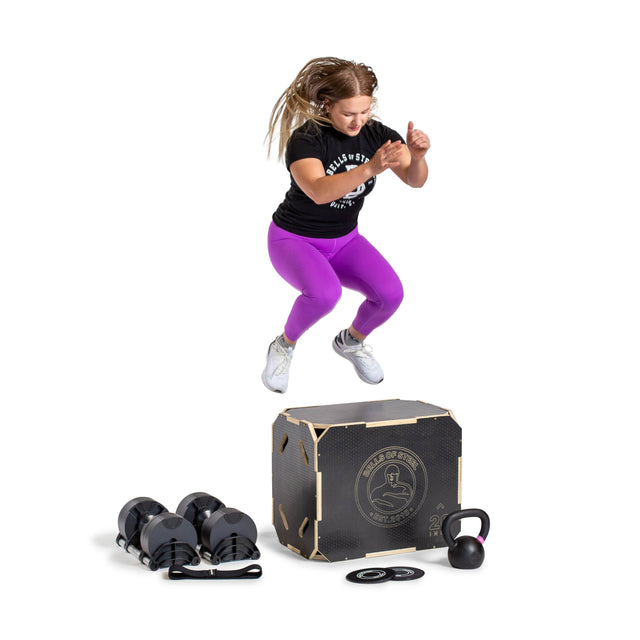 A woman in a black shirt and purple leggings jumps onto a plyo box, surrounded by dumbbells and Bells of Steel Strength Circuit Starter Kits—ideal for effective home workouts—all isolated on a white background.